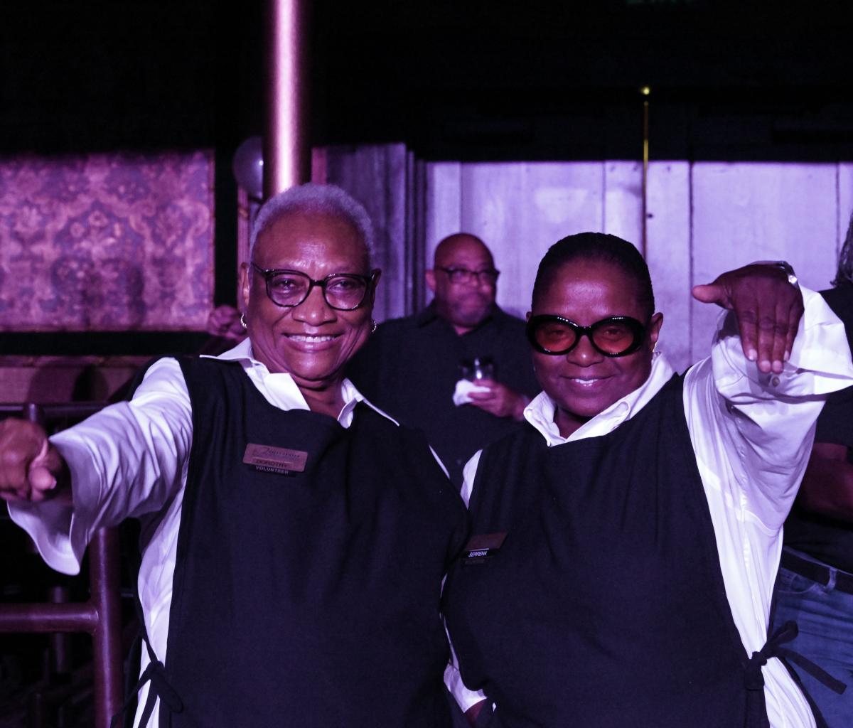 Two volunteers wearing black vests and name tags smile together inside a venue.